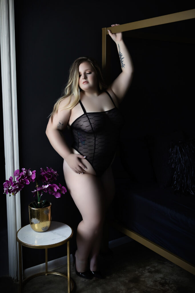 Plus size boudoir portrait of a woman standing next to the bed in edgy black one-piece lingerie, highlighting curves with bold and confident energy.