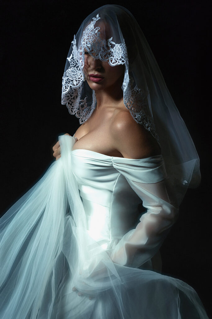 Fine art bridal portrait of a woman in a white wedding gown with a lace veil draped over her face against a dark studio backdrop.