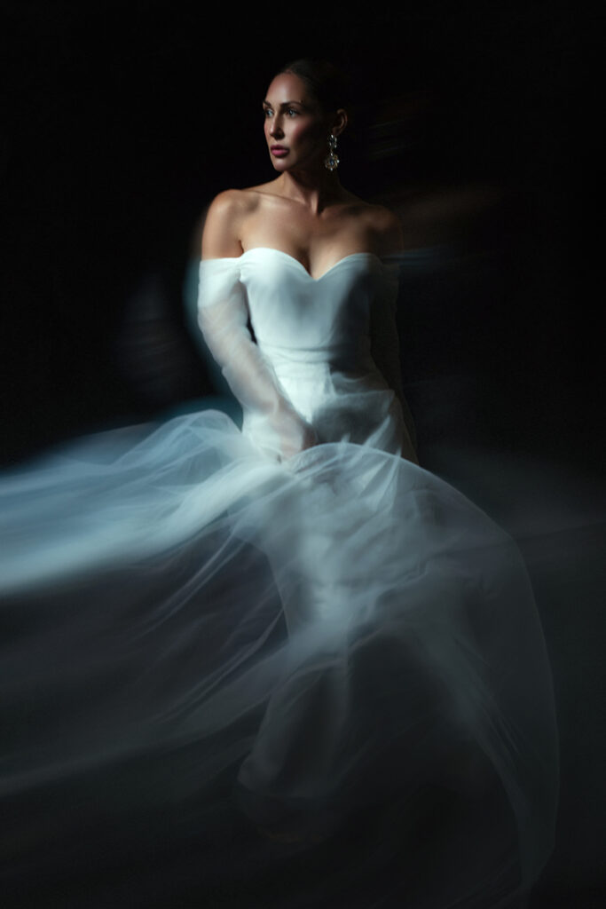 Artistic bridal portrait of a woman in a white wedding gown surrounded by soft, ethereal movement against a dark studio background.