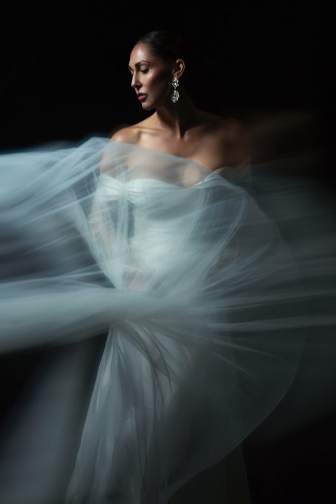 Ethereal bridal portrait of a woman in a white wedding gown surrounded by flowing translucent fabric and soft motion blur.