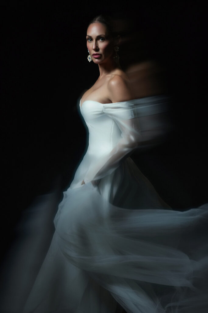 Editorial bridal portrait of a woman in a white wedding gown with flowing fabric and soft motion blur against a dramatic dark background.